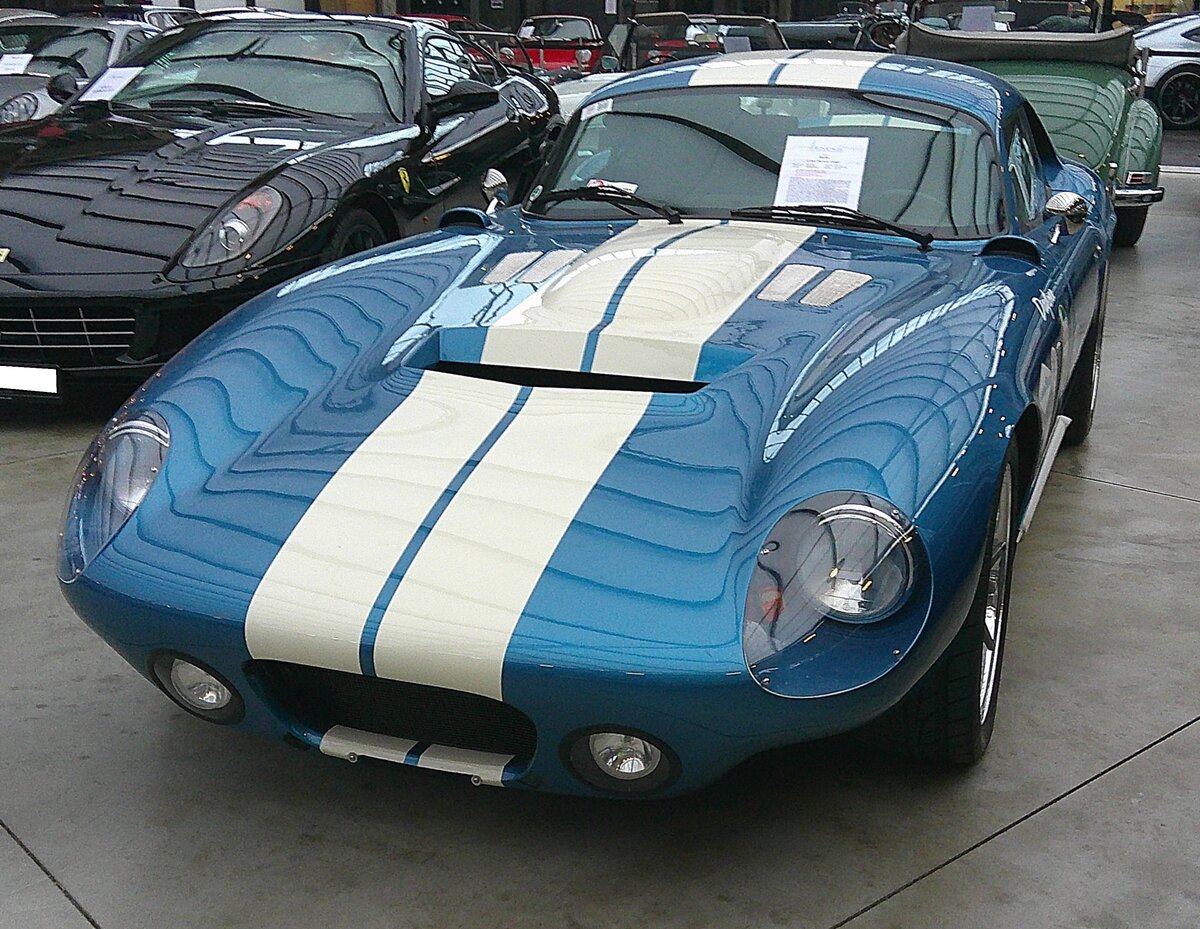 Shelby Cobra Daytona Coupe aus dem Jahr 1964. Das Shelby Cobra Daytona Coupe wurde konstruiert um die FIA-Sportwagenweltmeisterschaft zu gewinnen. Als einziger amerikanischer Hersteller gewann das Team des Texaners Carroll Shelby mit diesem
Modell die Meisterschaft im Jahr 1965. Peter Brock entwarf die glatte, aerodynamische Karosserie, die dem  Daytona  auf Hochgeschwindigkeitskursen in aller Welt den entscheidenden Vorteil verschaffte. In den 1960`er Jahren wurden nur sechs solcher Daytona-Coupes gebaut. Diese, CSX9000 genannte, Serie ist eine moderne Weiterentwicklung des Autos, der die Rennstrecken beherrschte. Die von den ursprünglichen Designern Peter Brock und Bob Negstad entworfenen und von Shelby American gebauten CSX9000
Continuation Daytona Coupés haben einen größeren Fahrgastraum als die ursprünglichen Rennwagen, was einen höheren Komfort ermöglicht. Außerdem verfügen sie über einen Rohrrahmen aus Baustahl. Dieser Wagen, mit der Fahrgestellnummer CSX9161, setzt den sportlichen Gedanken mit seiner Lackierung in der Farbkombination guardsman blue mit Streifen in wimbledon white fort. Der optimierte V8-Motor aus dem Hause Ford/USA hat einen Hubraum von 6997 cm³ und bringt satte 517 PS an die Hinterachse. Classic Remise Düsseldorf am 11.01.2025.