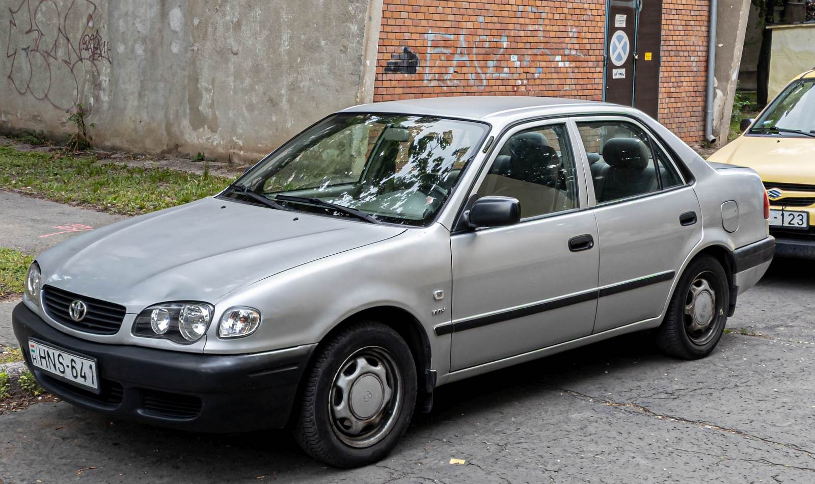 Hier ist ein Toyota Corolla E110 Stufenheck Facelift mit Basisausstattung (unlackierte Stoßtangen und Seitenspiegel) zu sehen. Die Aufnahme stammt von Mai, 2024.